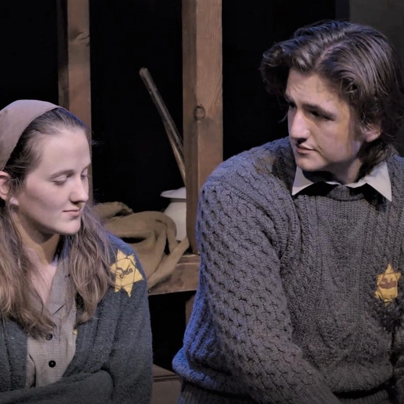 A girl and a boy, faintly smiling, wearing yellow stars proclaiming them as Jews on their sweaters, sit side-by-side in the cell of a concentration camp. 