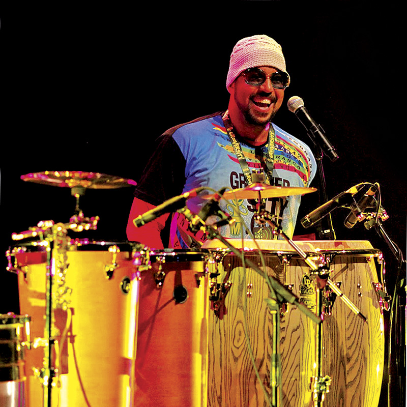 Dressed colorfully and wearing shades, Marcus Santos stands behind an array of Brazilian hand drums