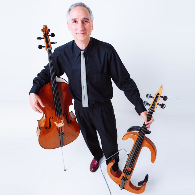 Gideon Freudmann with right arm around an acoustic cello and left hand holding an electric cello.
